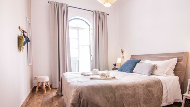 A neatly made bed with a wooden headboard, a small stool, and a nightstand with a lamp.