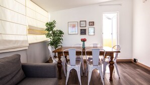 A dining area with a wooden table and white chairs, a potted plant, and framed pictures on the wall.