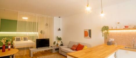 A modern living room with a sofa, a coffee table, and a TV. There is a kitchen area with a wooden countertop and a sink.