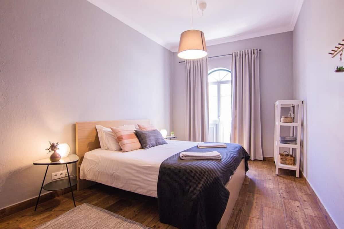 Un dormitorio con cama, mesita de noche, lámpara y una estantería con objetos.