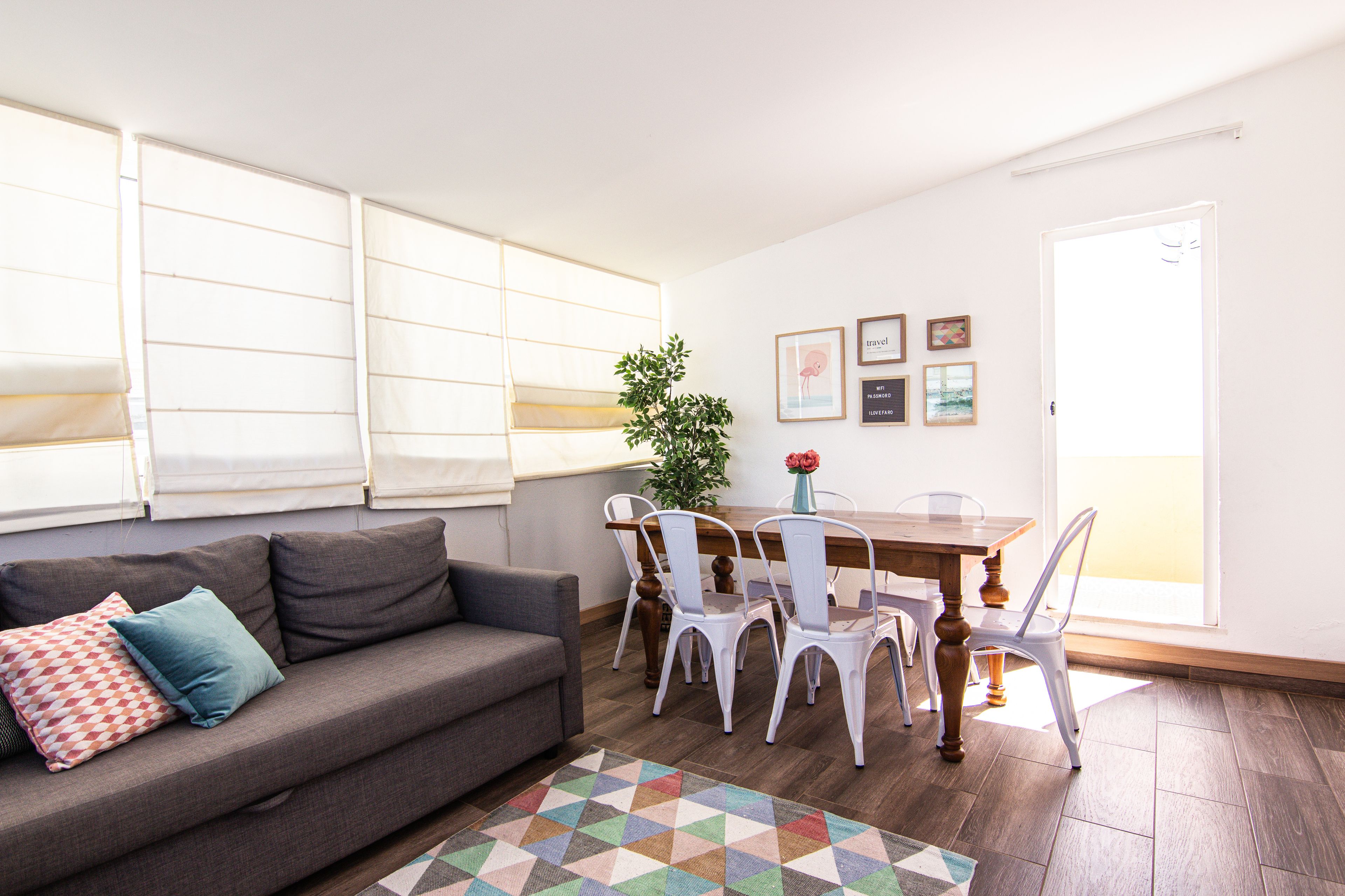 Een woonkamer met een bank, een eettafel met stoelen, een potplant en een raam met jaloezieën.