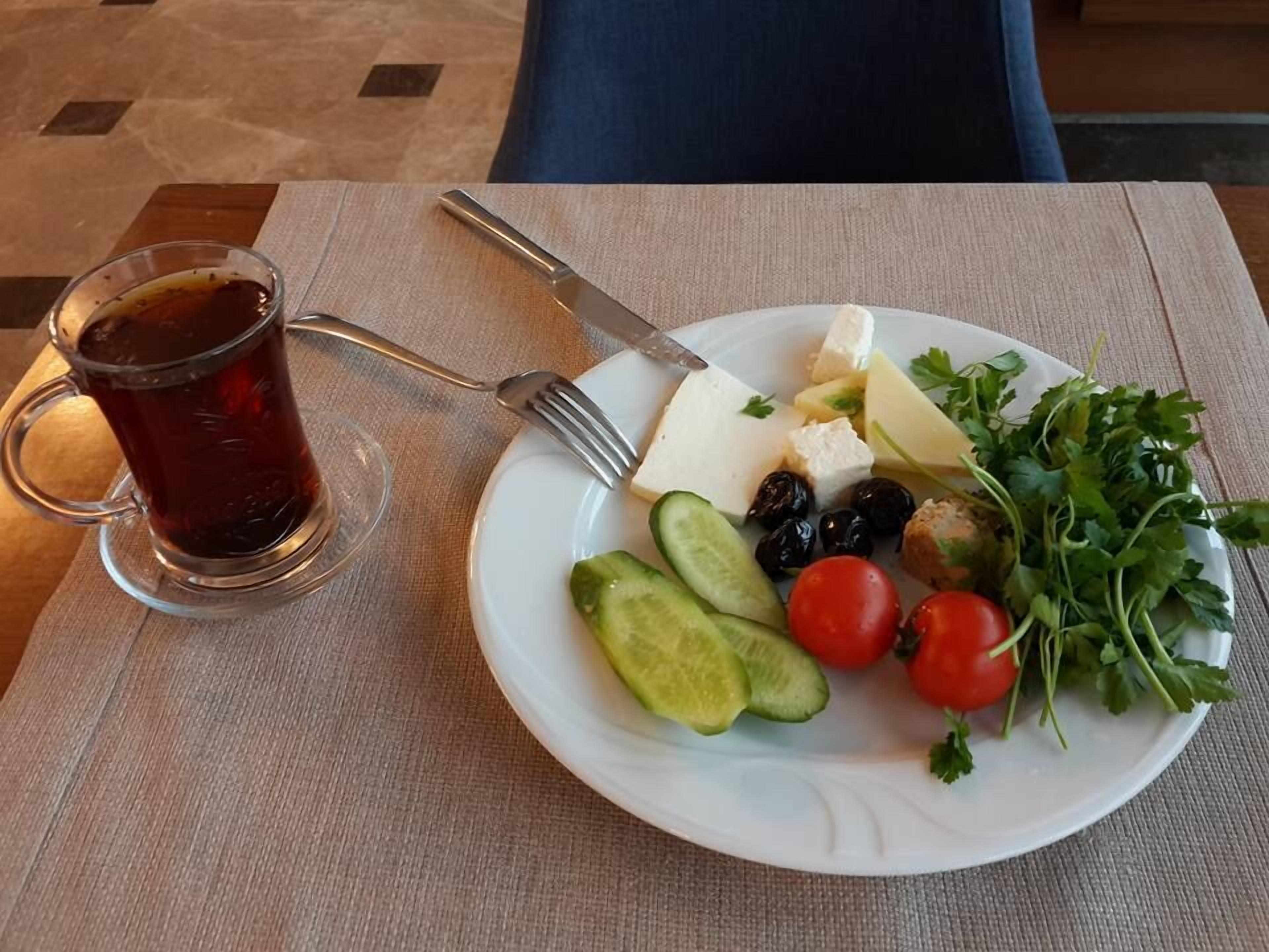 Un plato con queso, aceitunas, tomates, pepinos y perejil, acompañado de un vaso de té y cubiertos sobre una mesa.