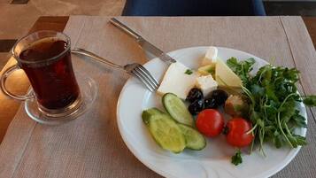 Un plato con queso, aceitunas, tomates, pepinos y perejil, acompañado de un vaso de té y cubiertos sobre una mesa.