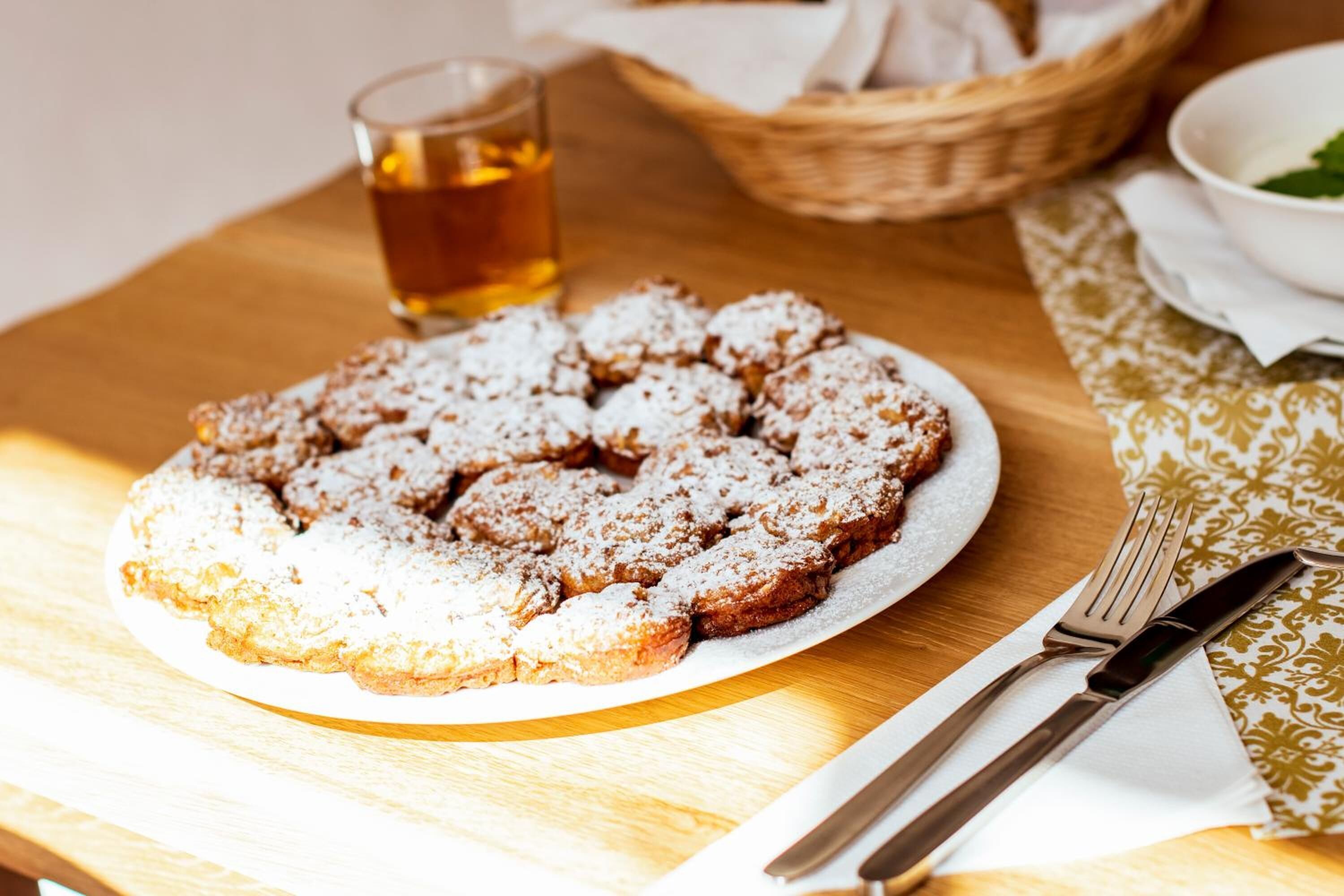 A plate of pastries dusted with powdered sugar on a wooden table with a glass of tea and cutlery.