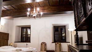 Habitación de hotel con techo de madera, una araña, dos ventanas, una cama, una mesa, dos sillas y un aparador.