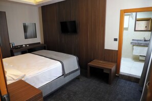 A hotel room with a bed, a television mounted on the wall, a small wooden bench, and a bathroom visible through an open door.