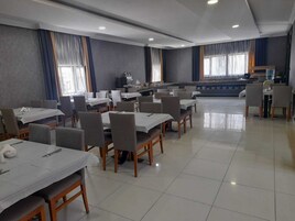 Un comedor amplio con varias mesas y sillas, una cocina al fondo y ventanas grandes con cortinas.