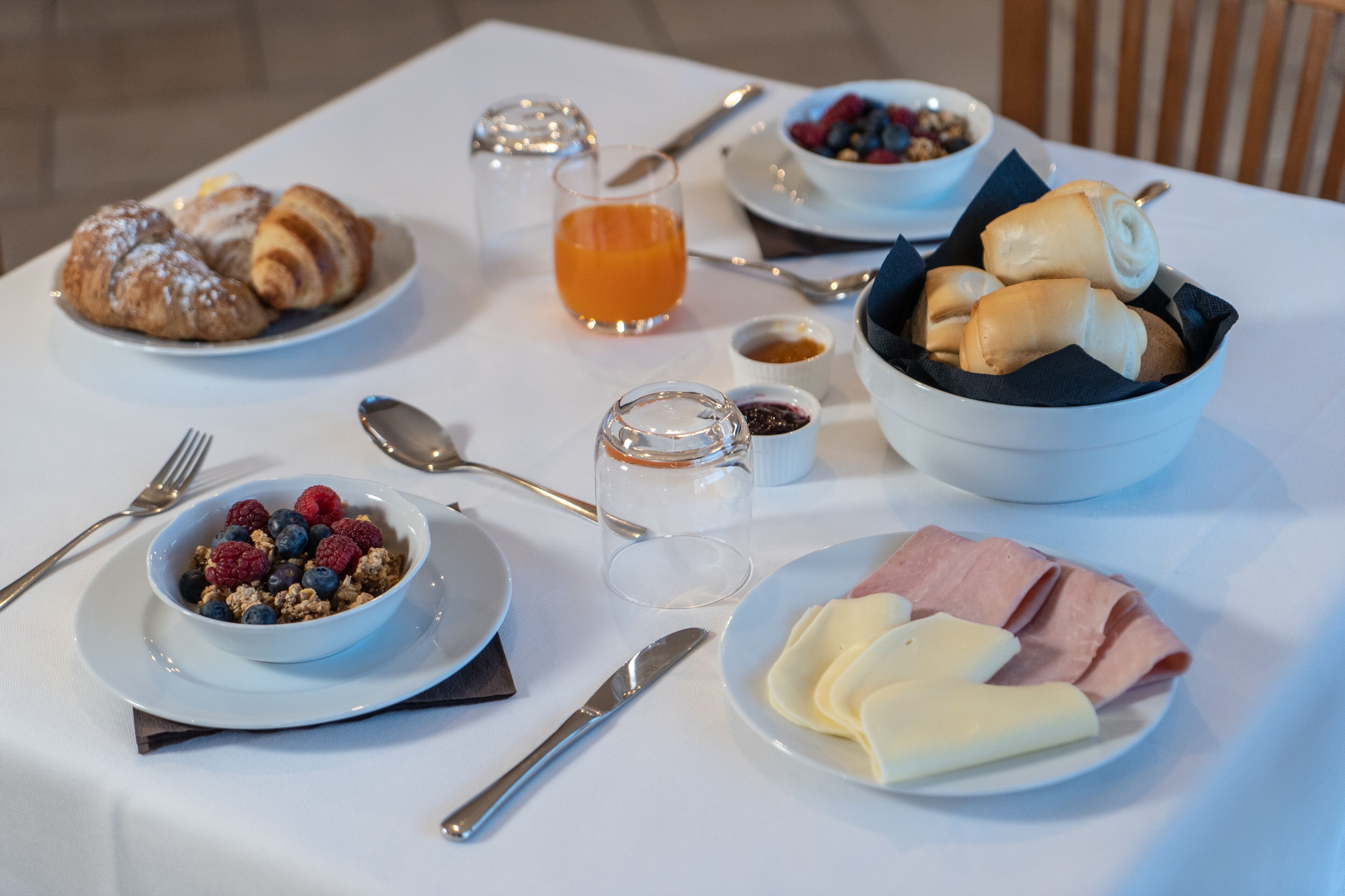 Una mesa de desayuno con cruasanes, un bol de bayas, un vaso de zumo de naranja y un plato de jamón y queso.