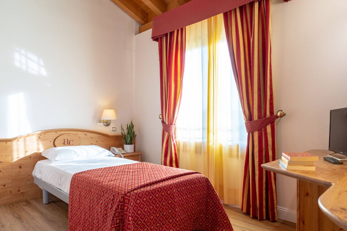 A hotel room with a wooden headboard, a bed with a red patterned bedspread, a bedside table with a lamp, a plant, and a TV on a wooden stand.
