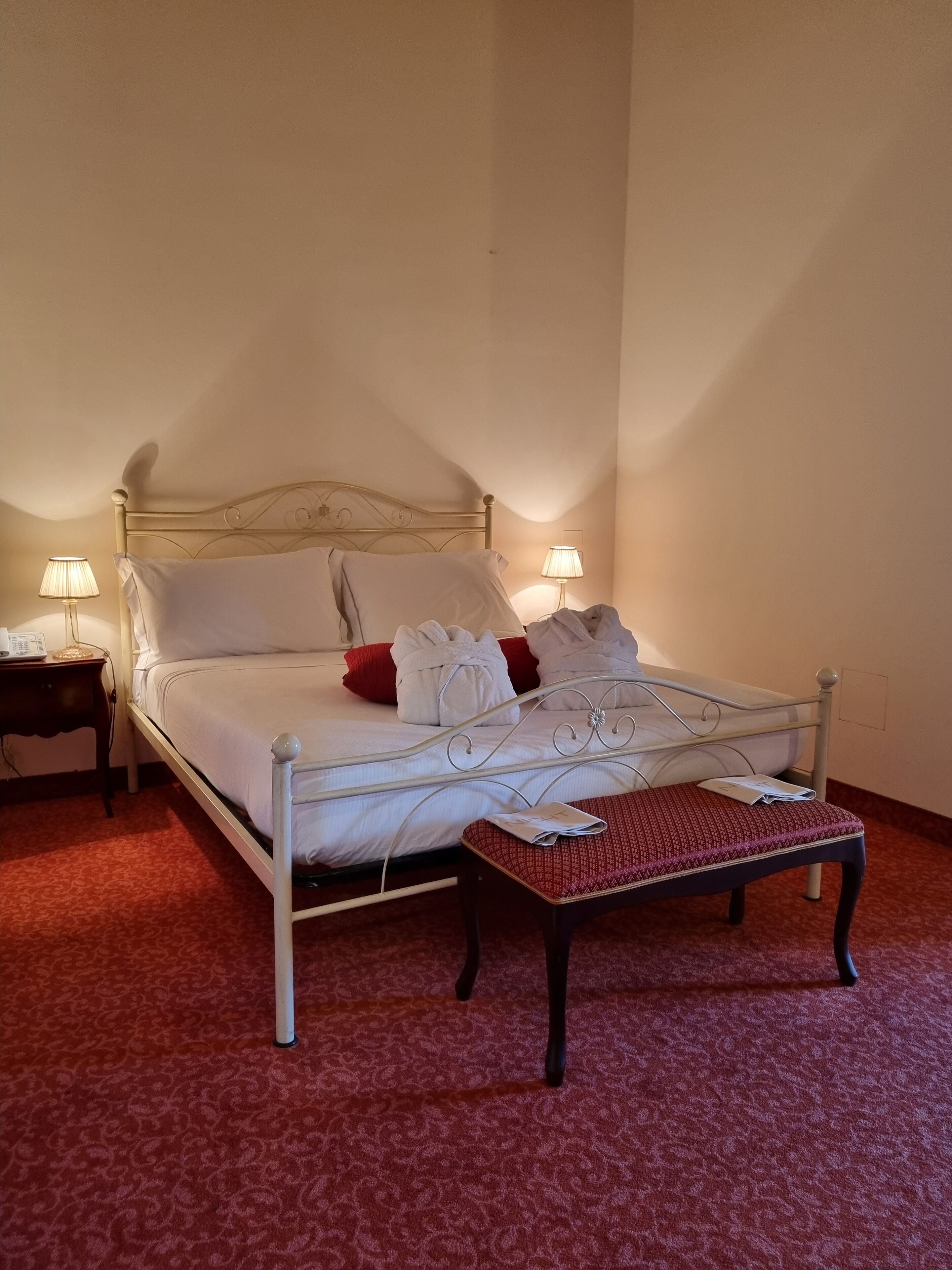 Una cama bien hecha con sábanas blancas y almohadas rojas, una mesita de noche con lámpara, y un otoman con estampado.