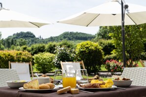 An outdoor dining table with breakfast items, surrounded by umbrellas and a scenic garden view. - Hotel Canova (Cavaso del Tomba)