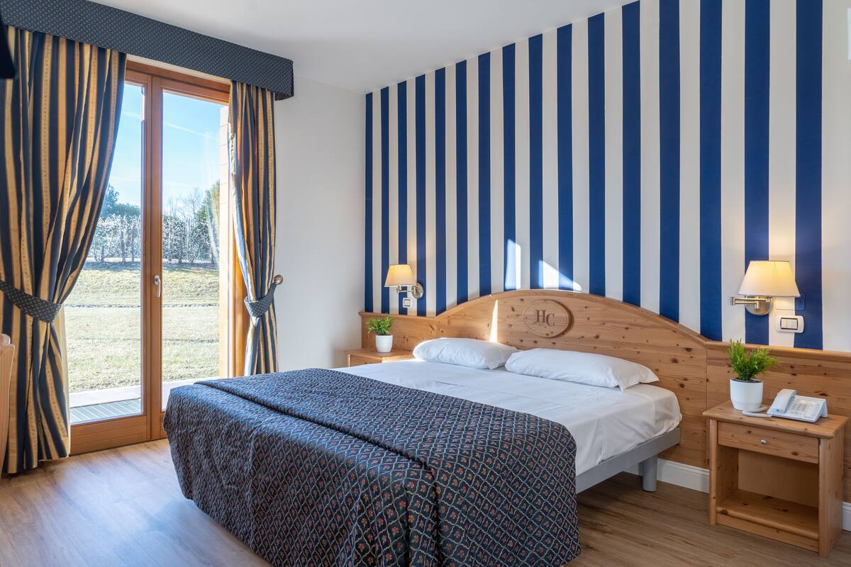 A bedroom with a large bed, wooden headboard, two bedside tables with lamps, a window with curtains, and a view of the outdoors.