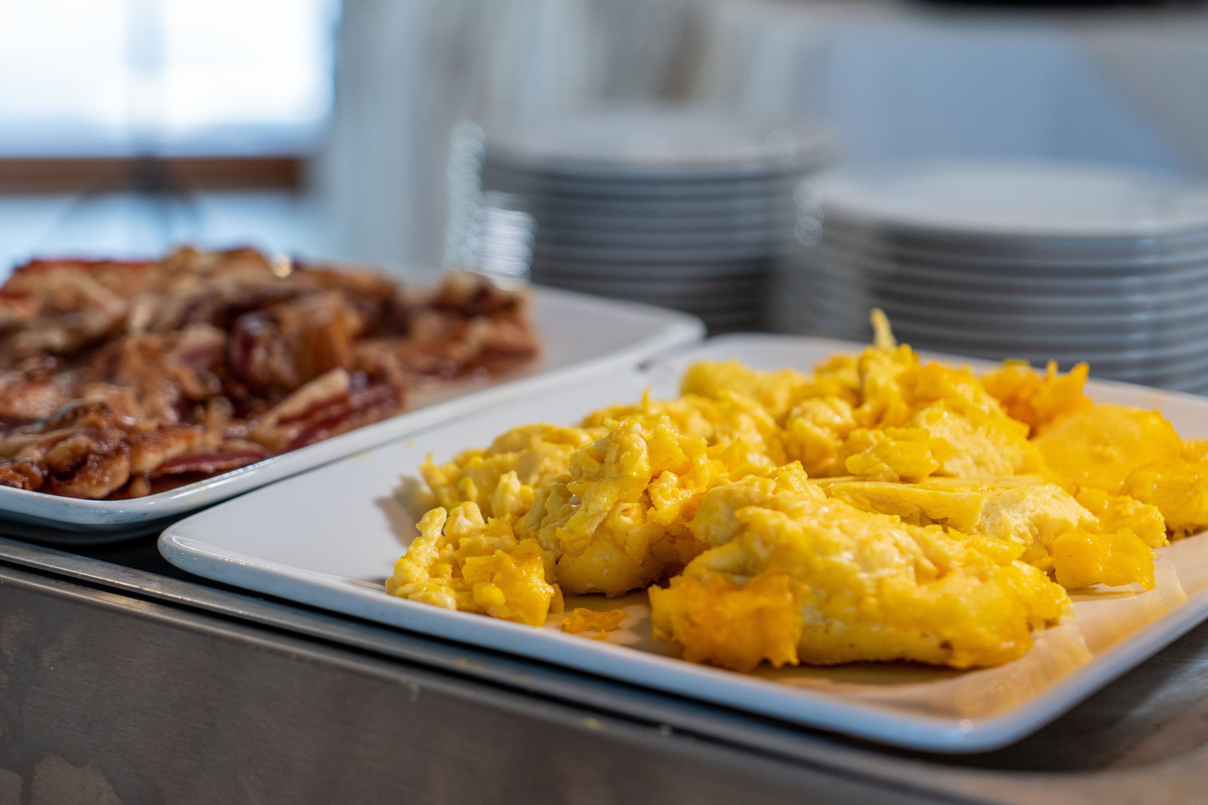 Un bufé de desayuno con huevos revueltos y tocino en platos blancos.