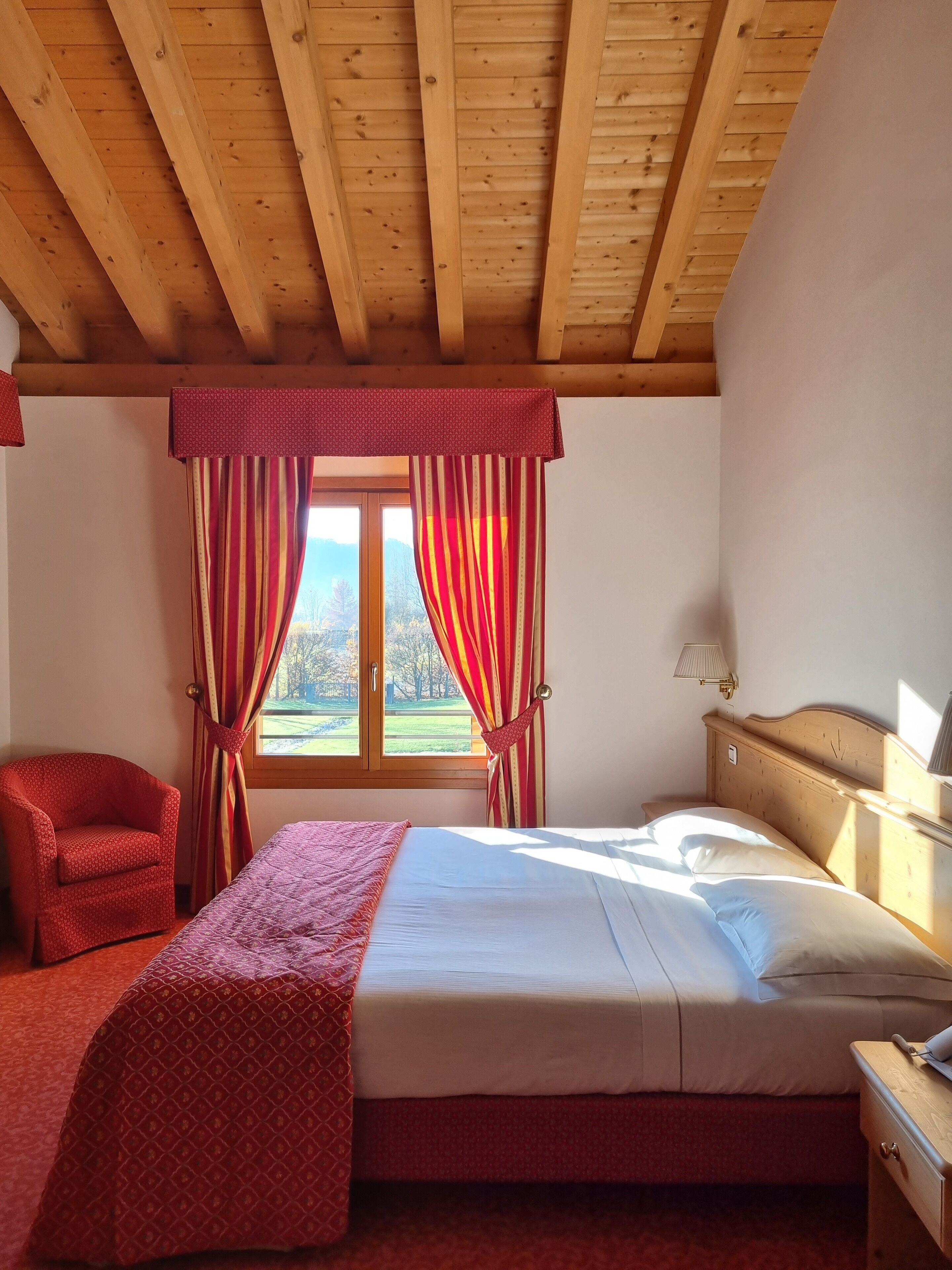 A bedroom with a wooden ceiling, a bed with white linens, a red armchair, and a window with curtains.