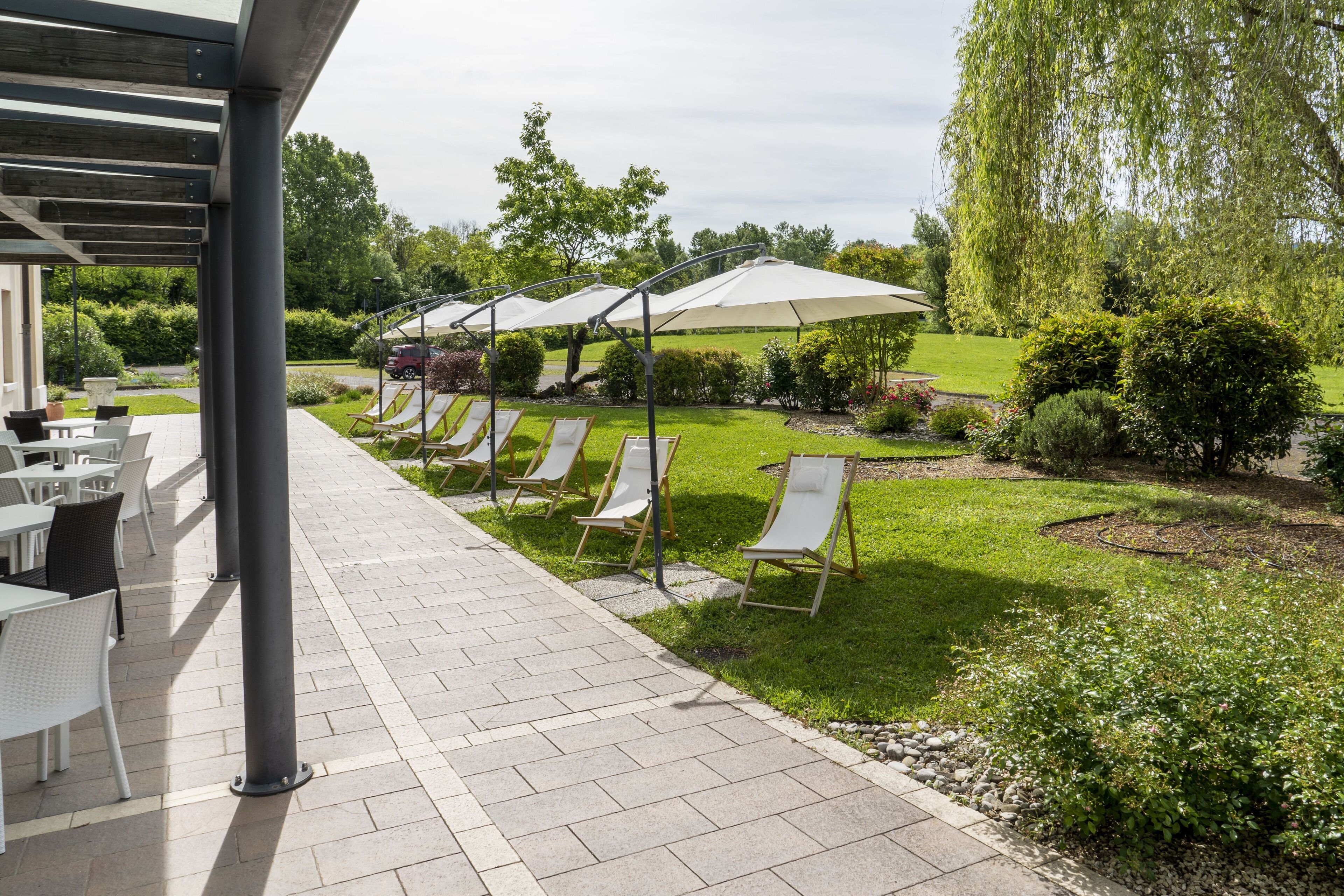 A covered outdoor seating area with lounge chairs and umbrellas, a paved walkway, and a well-maintained garden with hedges and trees.