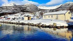 Paisaje nevado con un pequeño edificio cerca de un cuerpo de agua, reflejando las montañas circundantes.