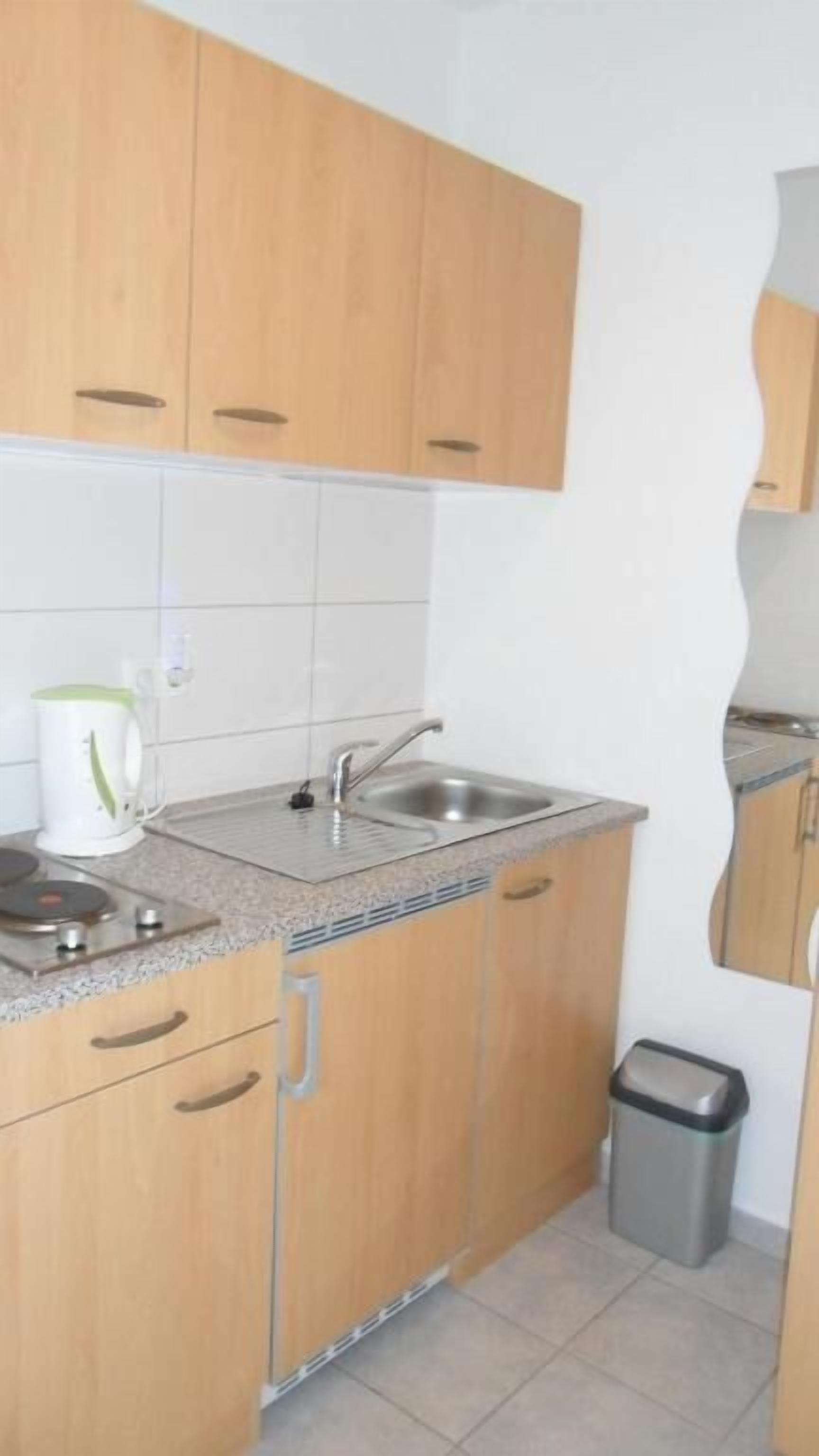 A small kitchen with wooden cabinets, a sink, a kettle, and a trash bin.