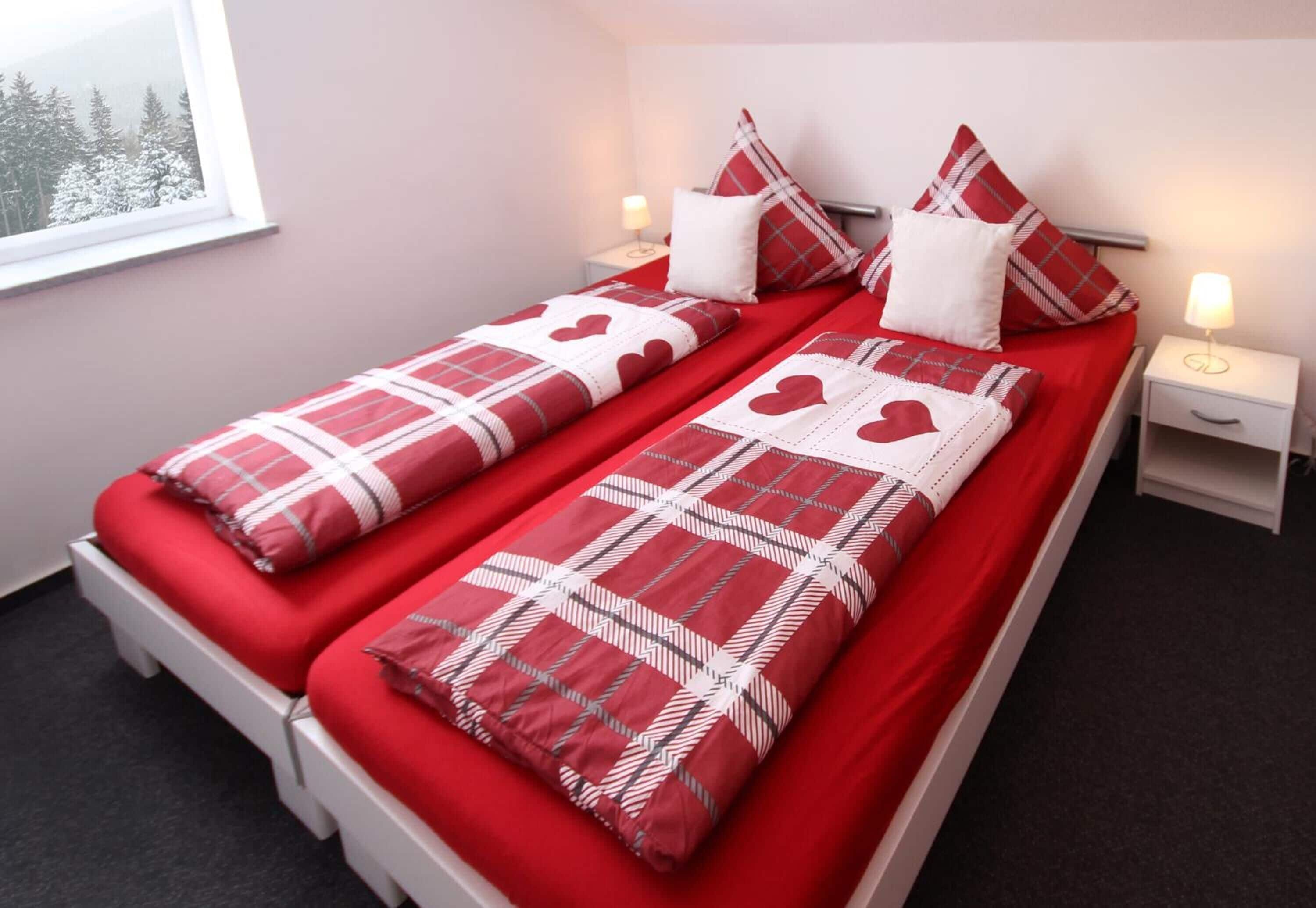 A double bed with red bedding and heart-patterned pillows, two bedside tables with lamps, and a window with a snowy view.
