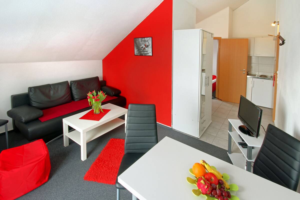 Un salon moderne avec un mur d’accent rouge, un canapé en cuir noir, une table basse blanche agrémentée d’un chemin de table rouge, et un coin repas avec une table et des chaises blanches.