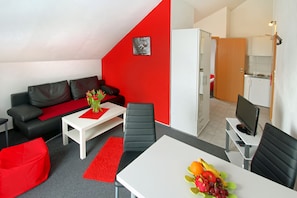 A modern living room with a red accent wall, a black leather sofa, a white coffee table with a red runner, and a dining area with a white table and chairs. - Appart-Hotel Harmonie (Winterberg)