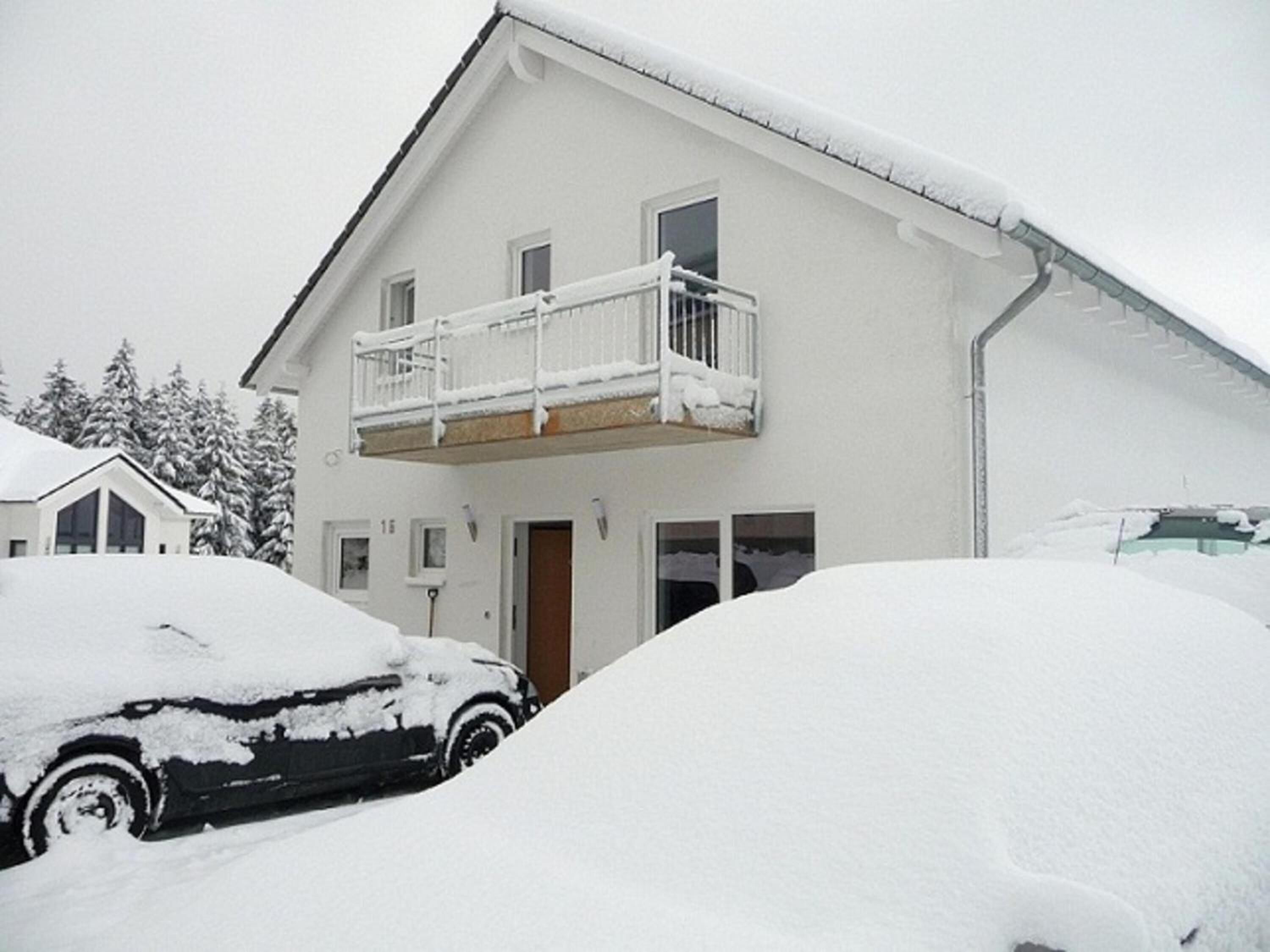 Una casa de dos pisos con balcón, rodeada de coches cubiertos de nieve.