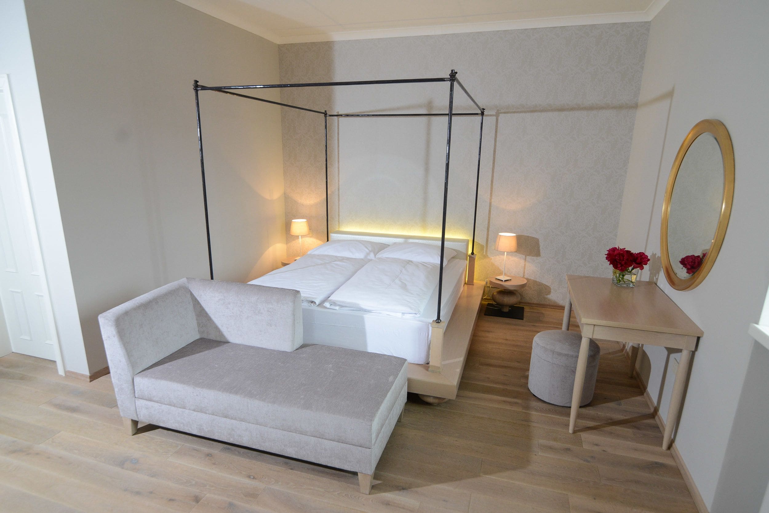 Une chambre à coucher moderne avec un lit à baldaquin, une table de chevet avec une lampe, une petite table ronde ornée d’un vase de fleurs et un grand miroir accroché au mur.