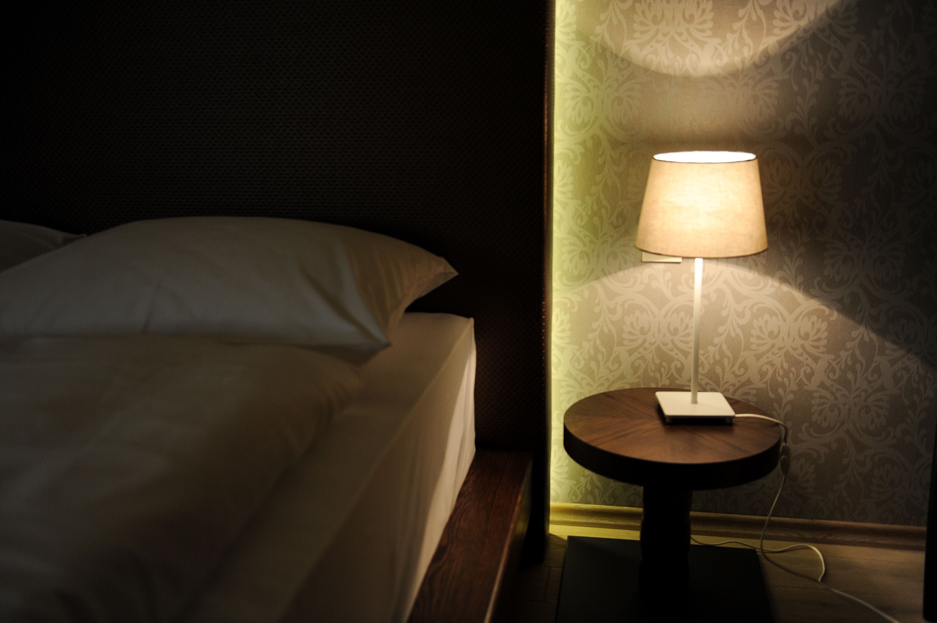 Een net opgemaakt bed met een wit kussen en een klein nachtkastje met een lamp en een stekkeradapter.