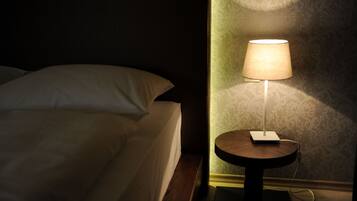Een net opgemaakt bed met een wit kussen en een klein nachtkastje met een lamp en een stekkeradapter.