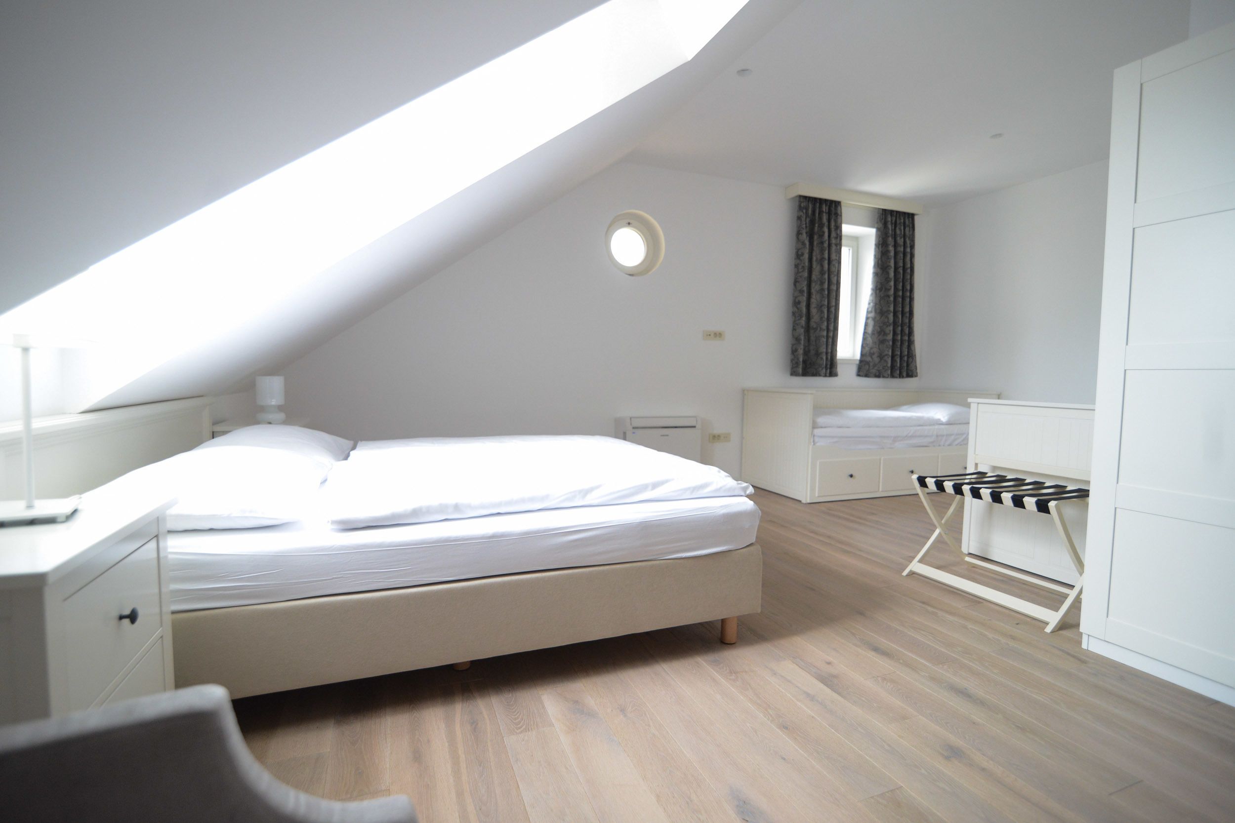 A bedroom with a sloped ceiling, a bed, a bedside table, a chair, a wardrobe, and a window with curtains.