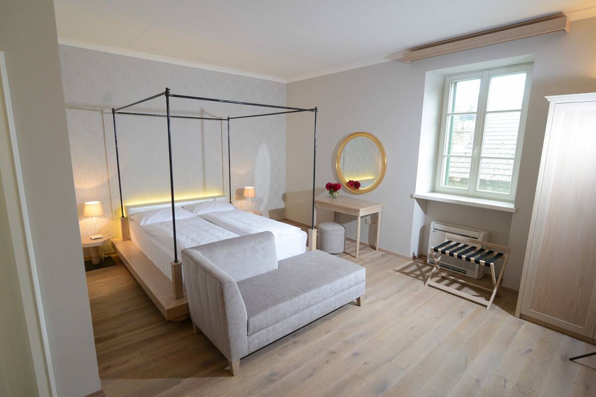 Une chambre à coucher avec un lit à baldaquin, une table de chevet avec une lampe, un petit miroir rond, une fenêtre avec des stores et un radiateur.