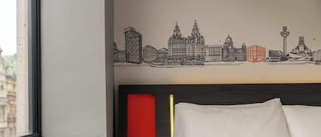 Quarto de hotel com um mural da paisagem urbana, uma cama com roupa de cama branca, uma parede de destaque vermelha e uma janela com vista para edifícios.