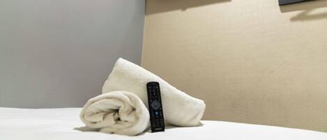 A bed with a rolled towel and a remote control, a television on the wall, and a coat rack.
