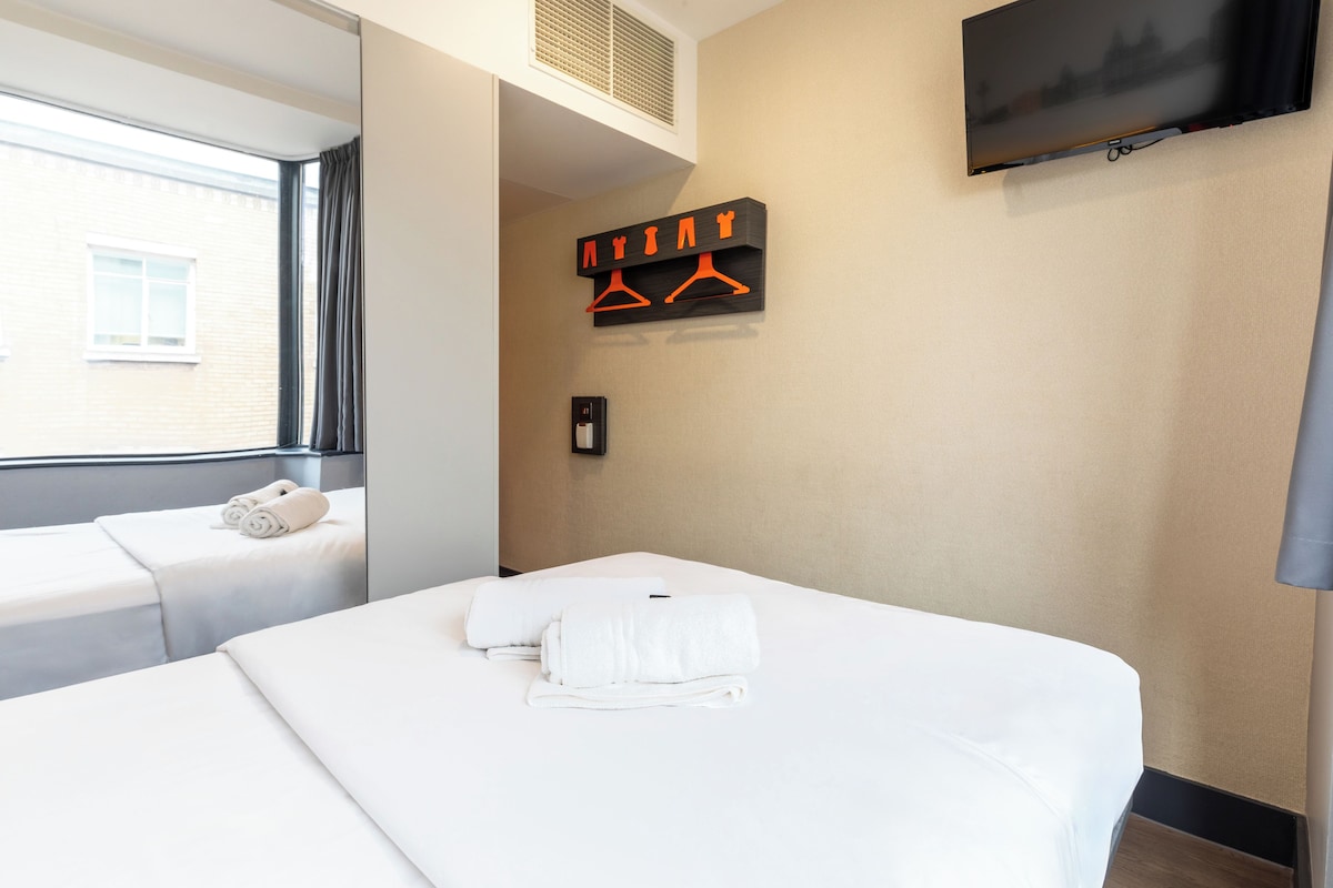 A hotel room with two beds, a TV mounted on the wall, and a window with a view of a building outside.