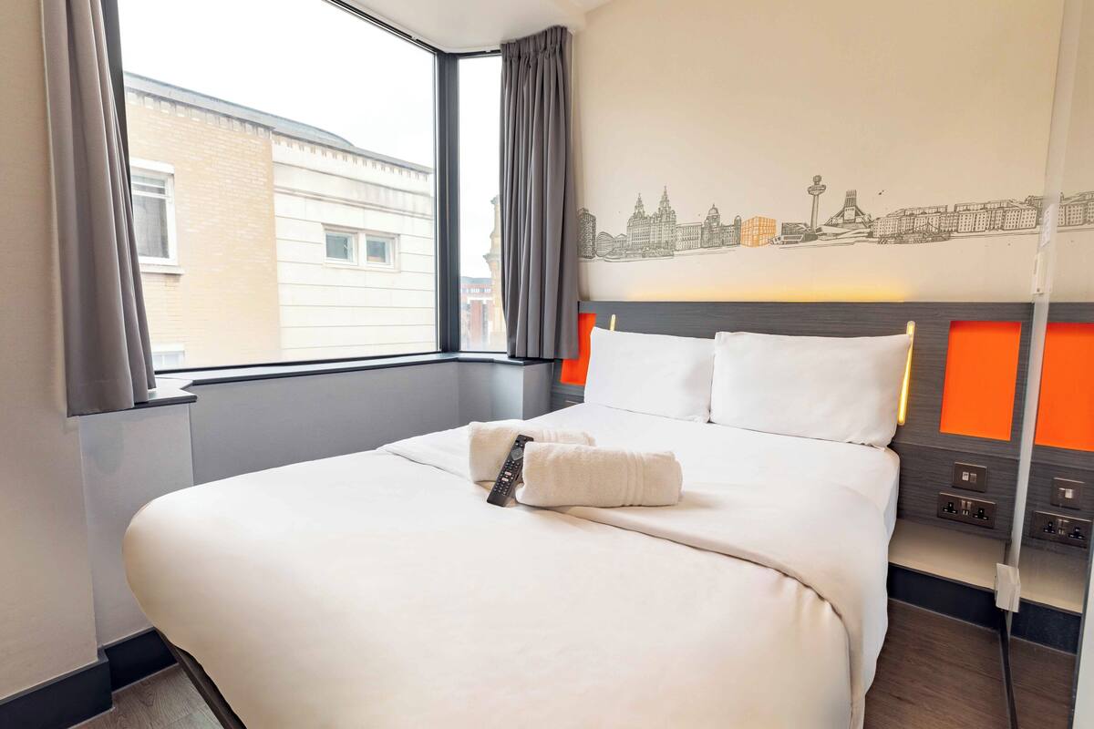 A hotel room with a large bed, a cityscape wall art, a window with curtains, and a bedside table with a lamp.