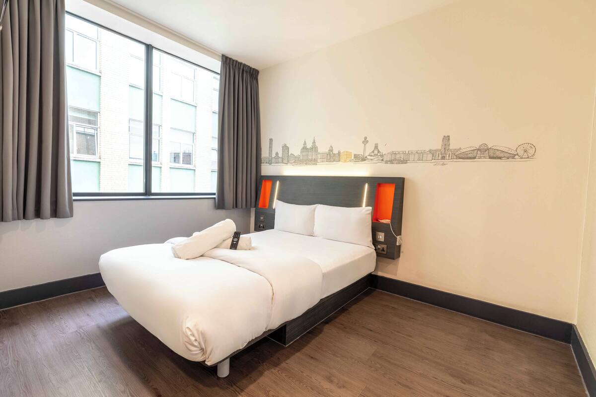 A hotel room with a bed, curtains, a cityscape wall art, and a window.