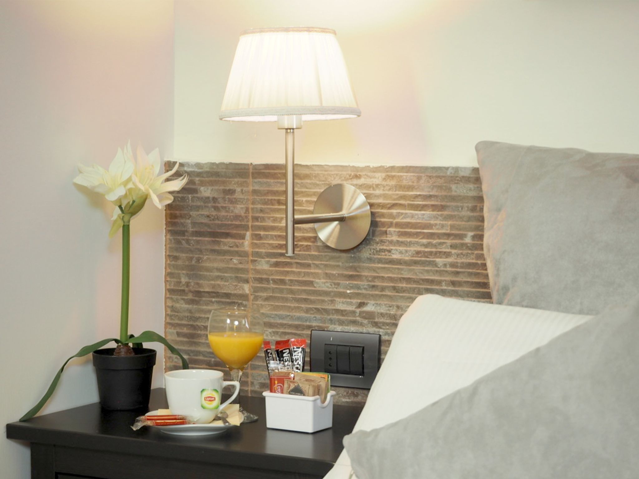 Een netjes ingerichte hotelkamer met een metselwerk-look muur, een moderne lamp, een wit kussen, een klein tafeltje met een potplant, een glas sinaasappelsap en een beker met een logo.