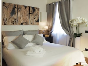 Un lit bien fait, avec des oreillers et des serviettes enroulées, une table de chevet avec une lampe, un mur orné d’une œuvre d’art à motifs de feuilles et une fenêtre avec des rideaux.