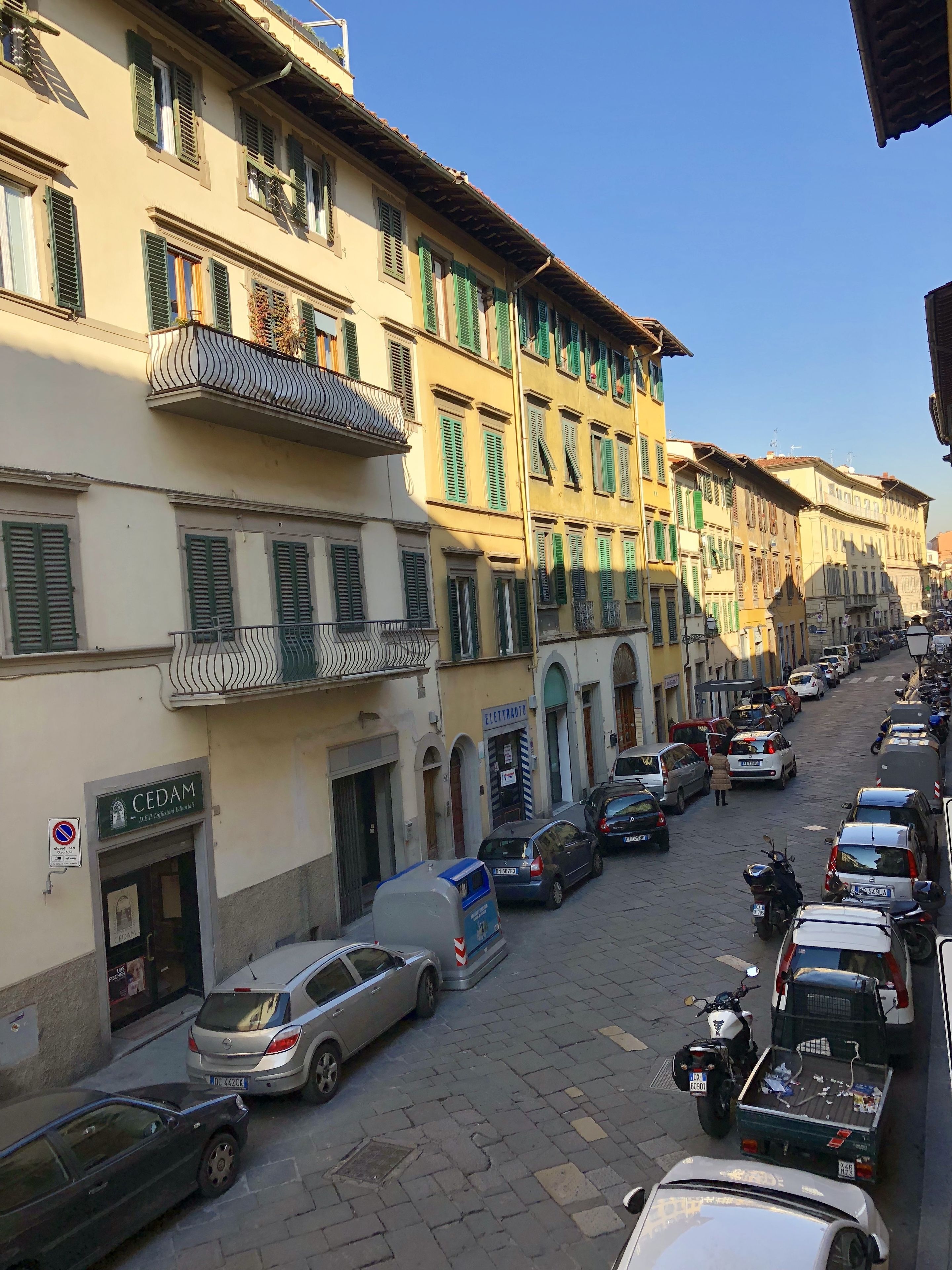A street lined with multi-story buildings, parked cars, and a few motorcycles.