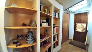 Corredor com uma estante de livros, objetos decorativos e uma porta com um cartaz.