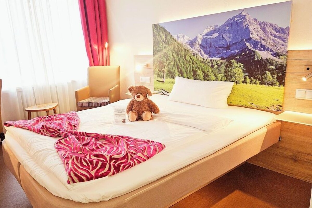 A hotel room with a bed, a teddy bear, a painting of mountains, a desk, and a chair.
