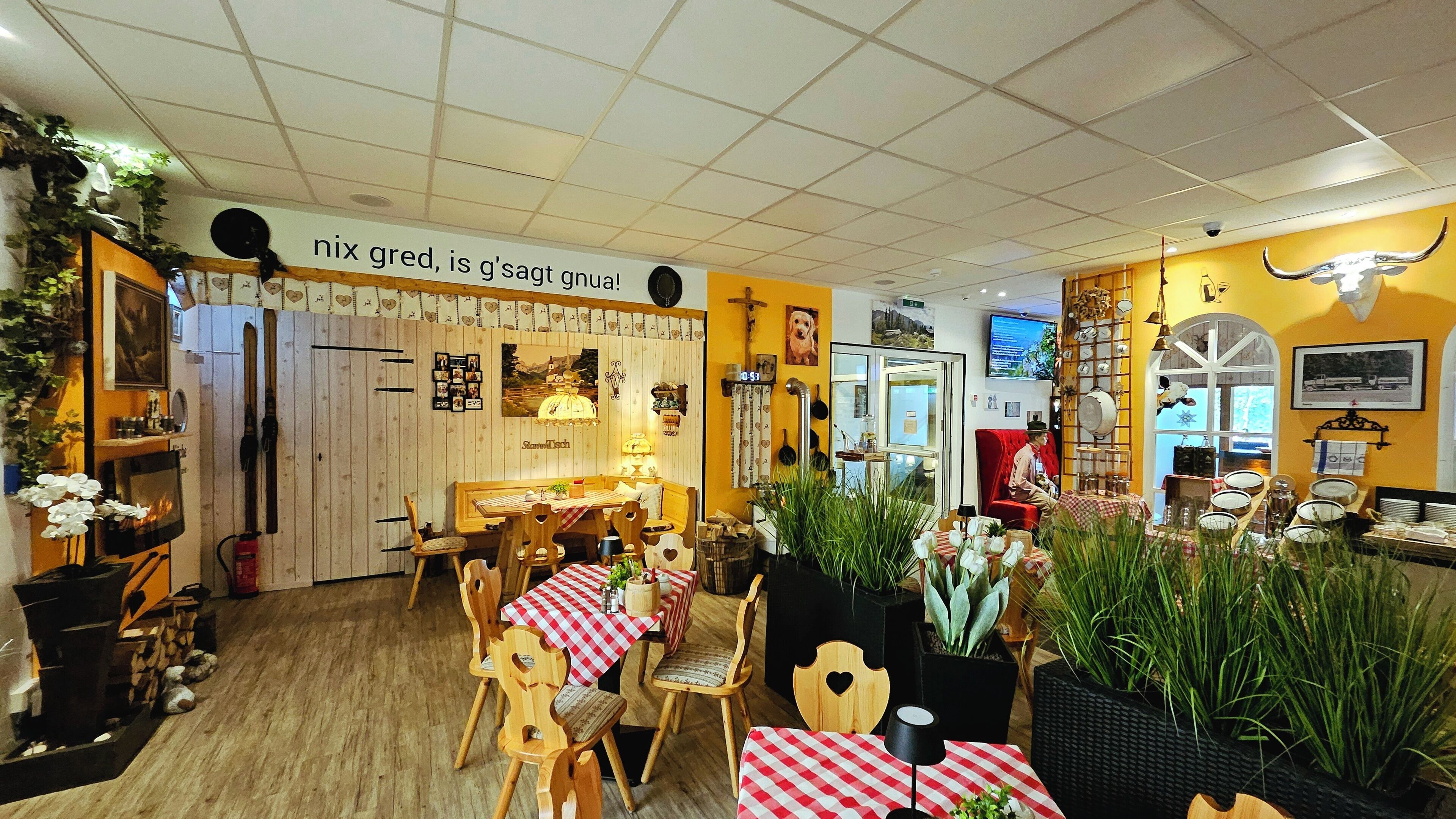 Interior de restaurante con mobiliario de madera, manteles a cuadros rojos y blancos, y una variedad de plantas.