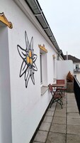 Fachada de edificio con pared blanca decorada con un mural de flores negras y amarillas, una ventana y un pequeño espacio al aire libre con una banca y una mesa.