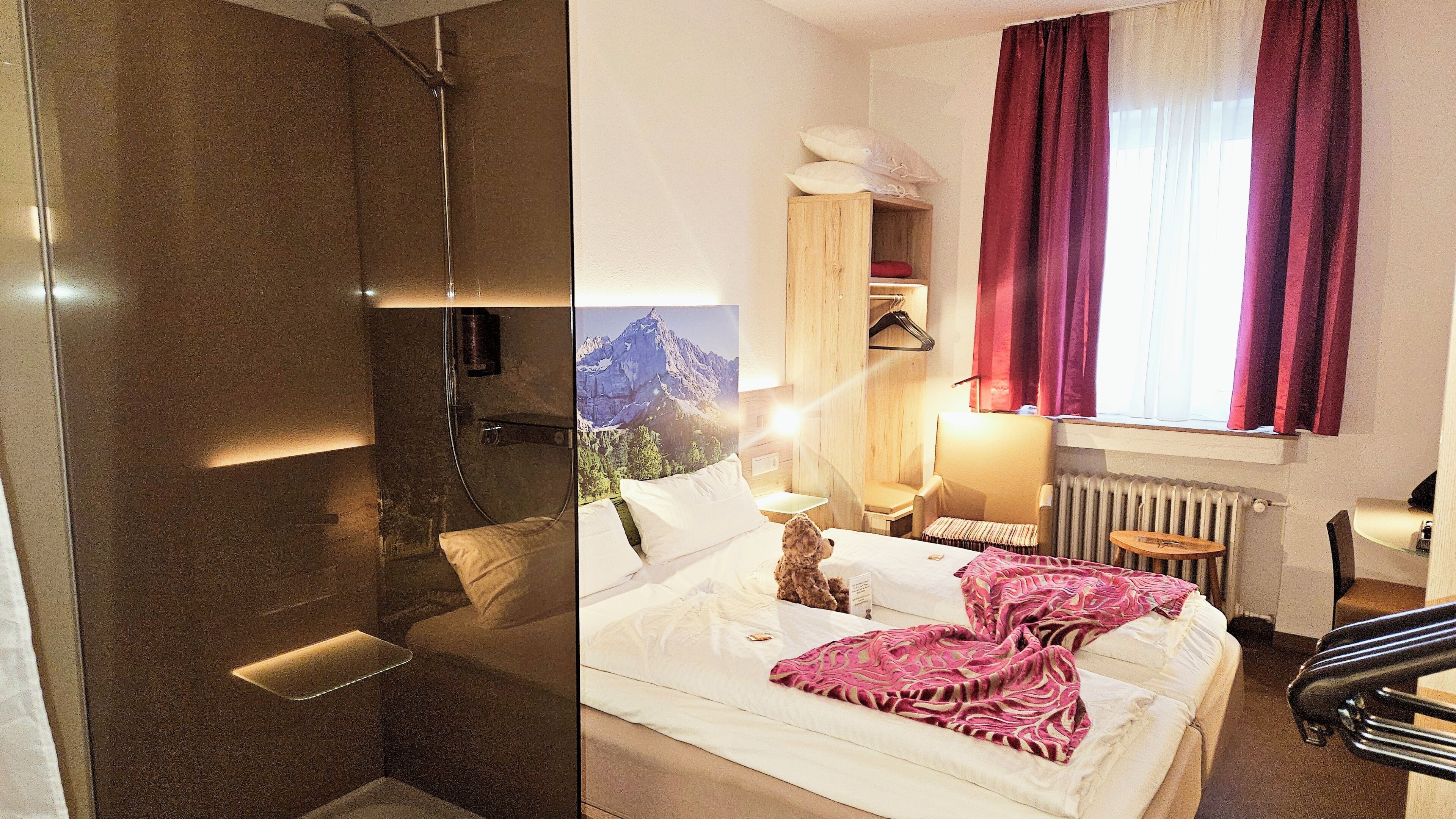 Quarto de hotel com chuveiro, uma cama com cobertor rosa, uma cadeira, uma mesa e uma janela com cortinas.