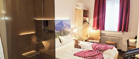 Ein Hotelzimmer mit Dusche, einem Bett mit rosa Decke, einem Sessel, einem Tisch und einem Fenster mit Vorhängen.