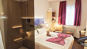 Habitación de hotel con ducha, cama con manta rosa, silla, mesa y ventana con cortinas.