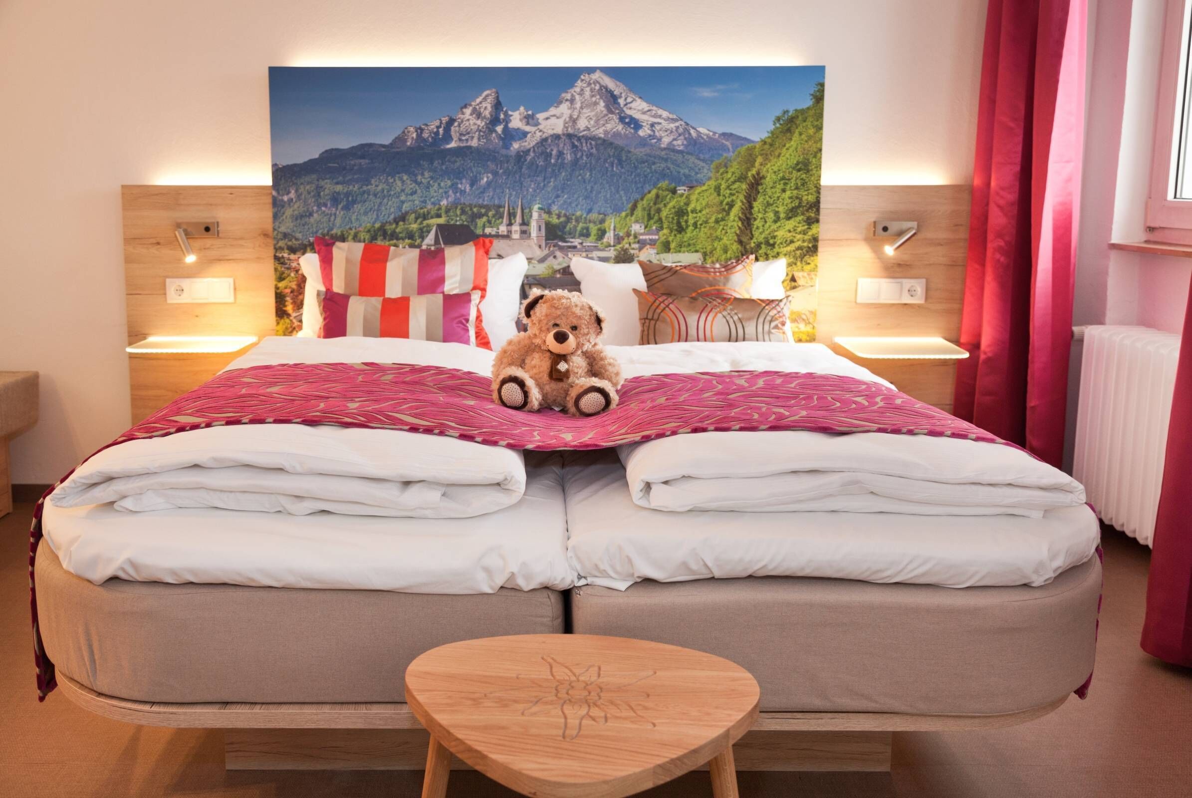 Ein Doppelbett mit Teddybär, zwei Nachttische mit Lampen, ein Holz-Kopfteil und Blick auf Berge durch ein großes Fenster.