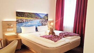 Habitación de hotel con cama, escritorio, silla, un cuadro de un paisaje de montaña y un osito de peluche en la cama.