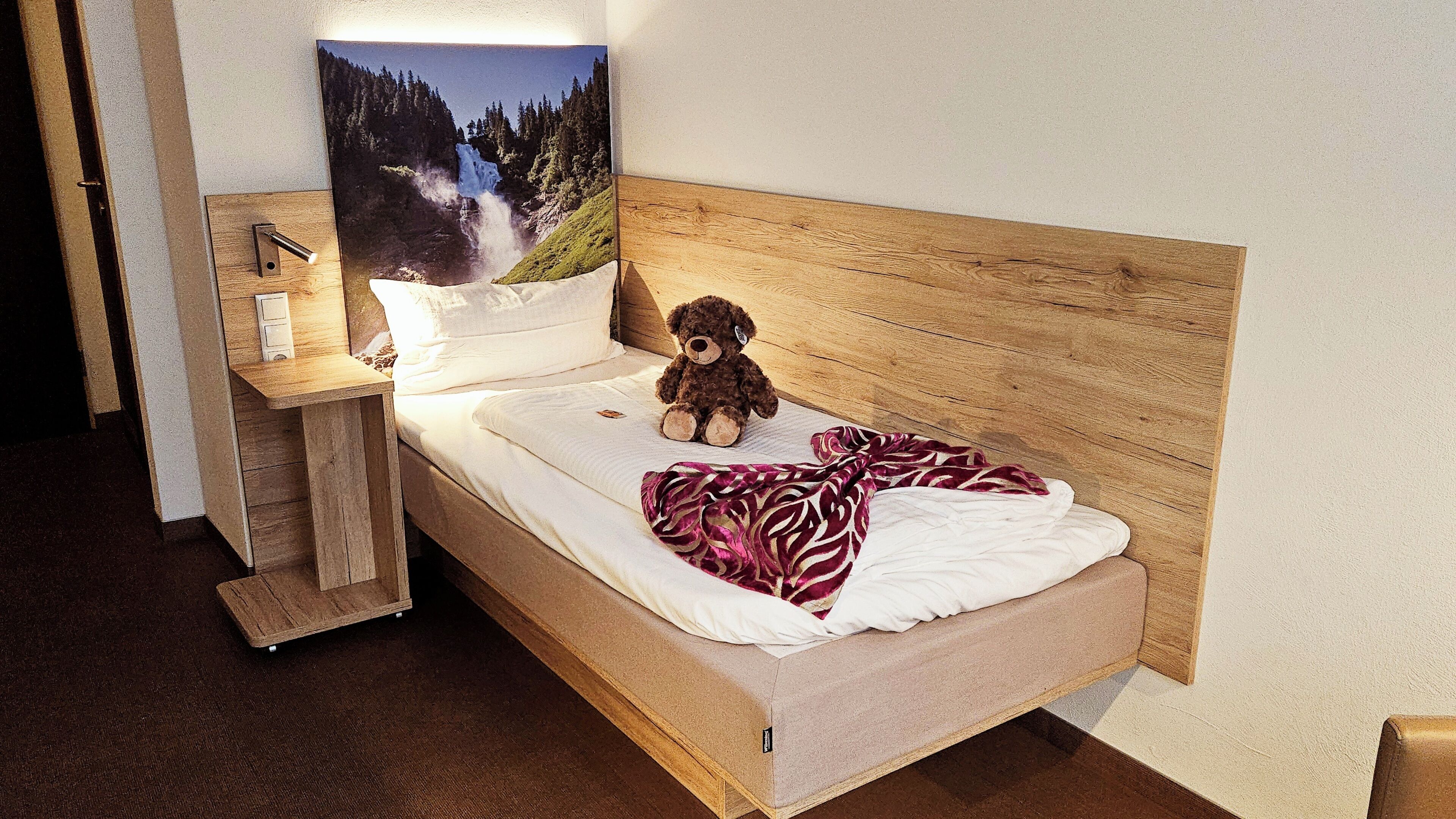 A single bed with a wooden headboard, a teddy bear, a blanket, and a nightstand with a lamp.