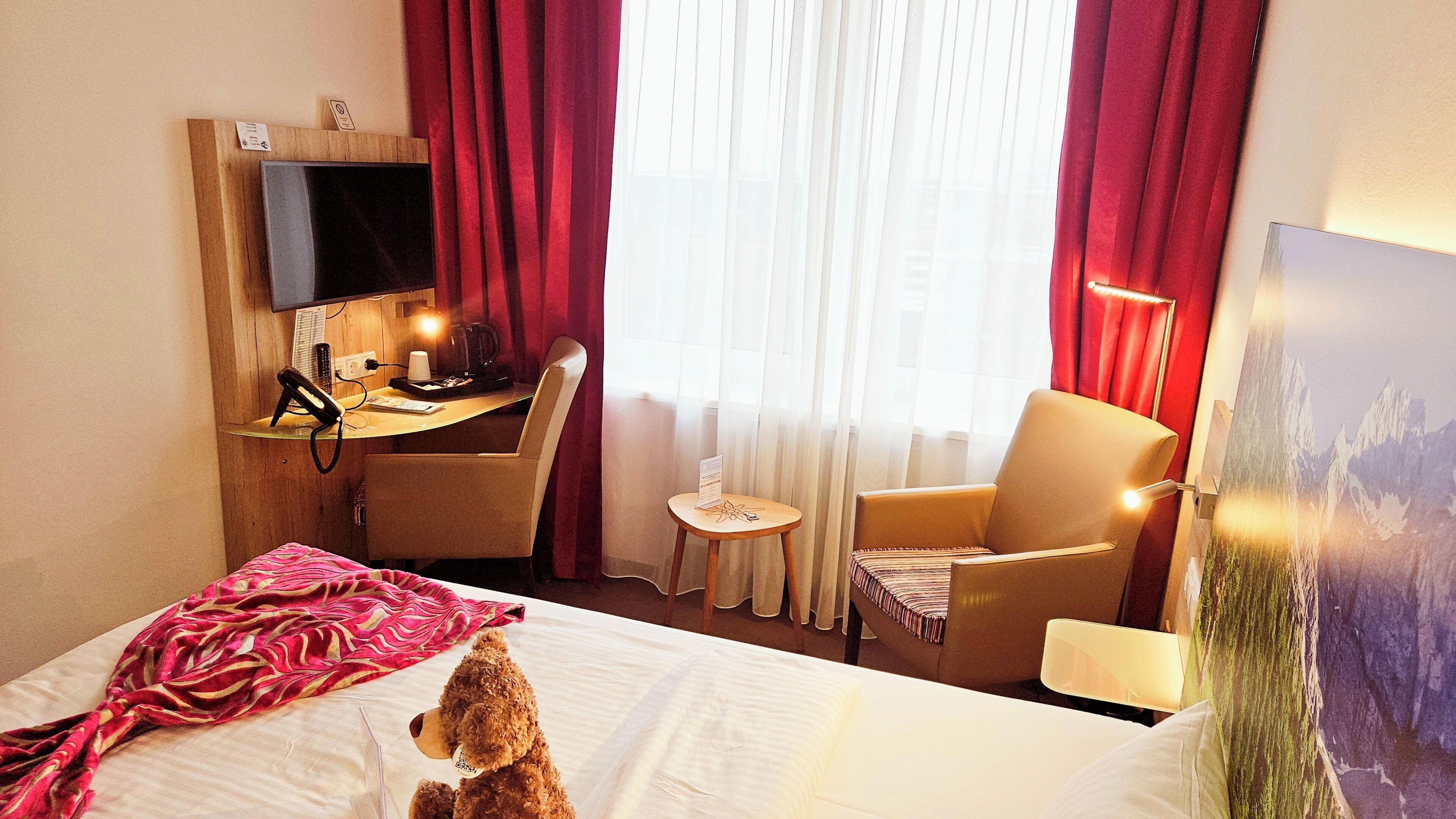 Una habitación de hotel con cama, escritorio con computadora, silla, una mesita y televisor montado en la pared.