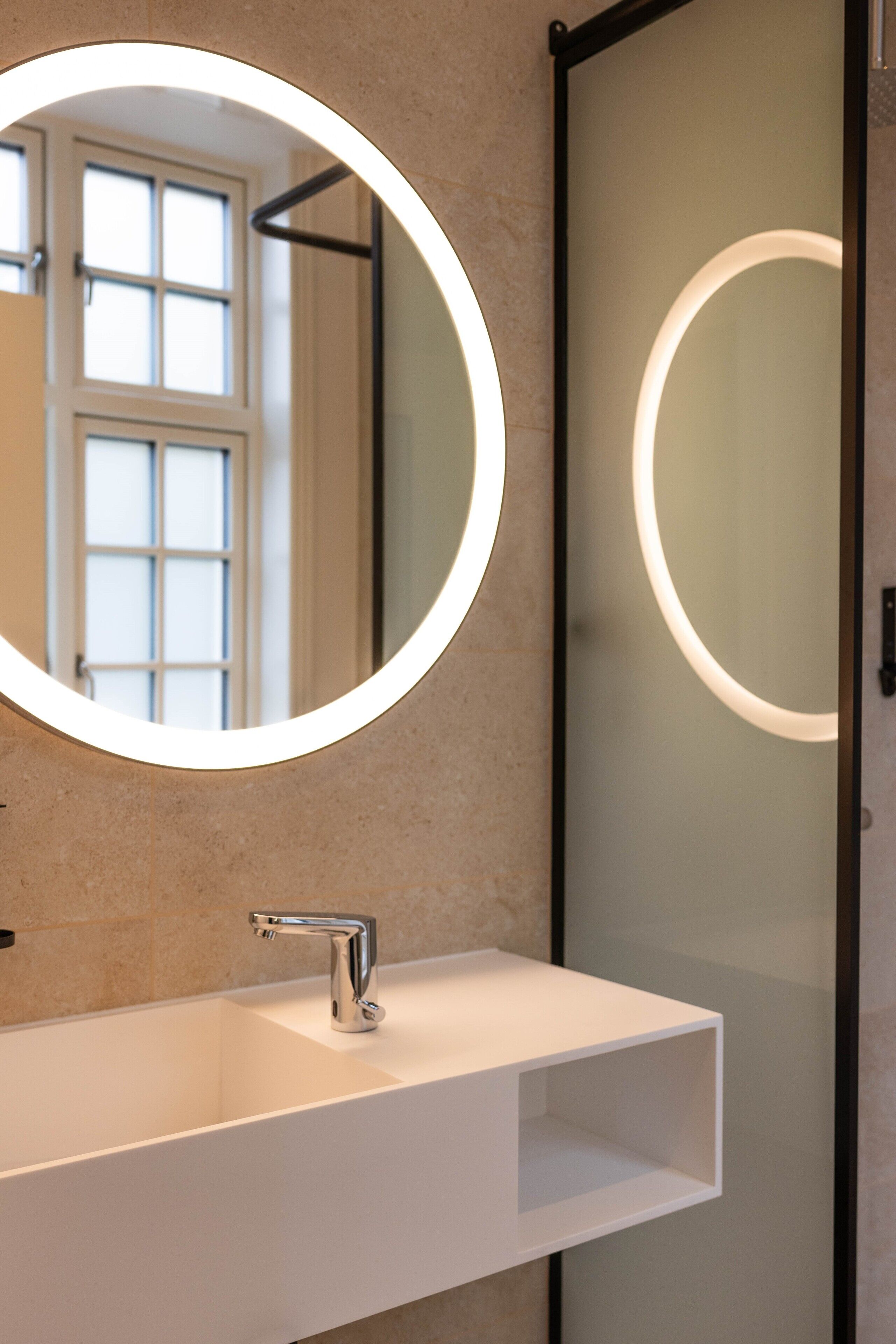 A bathroom with a white sink, a round mirror with a light, and a frosted glass door.
